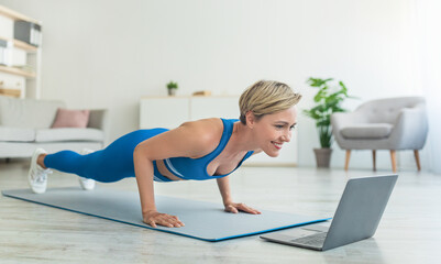 Fototapeta na wymiar Cheerful woman doing plank on mat with laptop at home