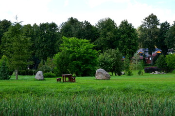 View of a beautiful and well maintained public park including a number of rocks, boulders, stones, benches, and paths located next to a dense forest or moor, as well as to a river bank seen in summer