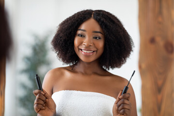 Portrait of pretty young Afro woman holding mascara in front of mirror, using decorative makeup at...