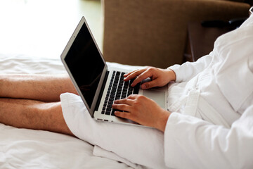 a freelancer on vacation works at a laptop lying on a bed in a white coat. Close-up, no face. A man's hands on a computer keyboard. Work during the holidays in the hotel room