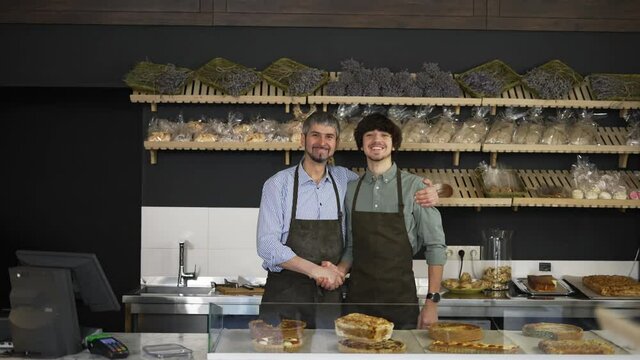 Close up portrait of happy successful family business owners handshaking in their bakery store looking at camera and smiling. Father and son