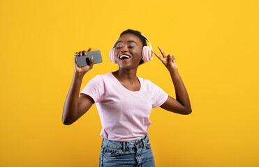 Joyful african american lady singing into smartphone and listening music via wireless headphones...
