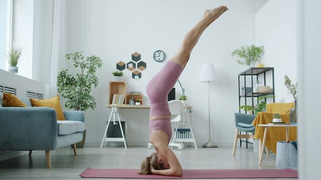 Portrait of attractive girl in sportswear doing headstand enjoying yoga exercises indoors at home. Active lifestyle and healthy youth concept.