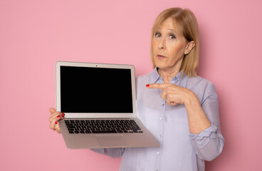 Senior woman showing laptop computer screen standing isolated on pink background