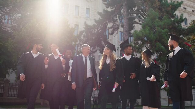 In the college garden group of multiracial students graduates at the end of the graduation in front of the camera walking all together with the college principal and smiling large feeling very excited