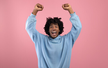 Triumphant black teen guy lifting arms above his head, shouting in excitement on pink studio...