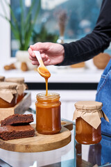 a hand with a spoon on which viscous homemade caramel flows into a jar against the background of other packed jars