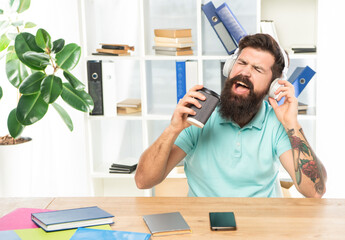 Happy businessman singing to takeaway cup listening to song in headphones in office, music