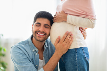 Delighted eastern man embracing tummy of his pregnant wife, listening baby kicks, tenderly touching belly of his spouse