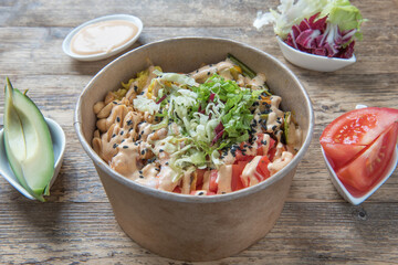 Healthy and delicious vegan poke bowl with zucchini, mixed salad, tomatoes, salad tomatoes, carrots, avocado, brown rice, black rice, cous cous, chickpea hummus, lentils on wooden table surrounded by 