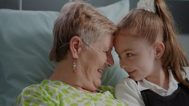 Close up of senior woman with illness enjoying visit from litte girl in hospital ward bed. Aged patient with sickness having fun with niece visiting to give support and assistance.
