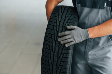 Man in uniform is working in the autosalon at daytime. Holding tire in hands