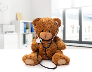 medicine, healthcare and childhood concept - teddy bear toy with stethoscope over medical office at hospital background