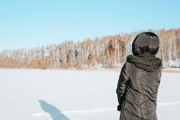 Fototapeta na wymiar Back view of alone woman in hood and jacket standing on sunny frosty day in nature against backdrop of snowy forest and looking at winter landscape, copy space