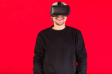 Man in cap dressed in black wearing Virtual Reality glasses, smiling, on red background. Technology, VR, computing and hobbies concept.