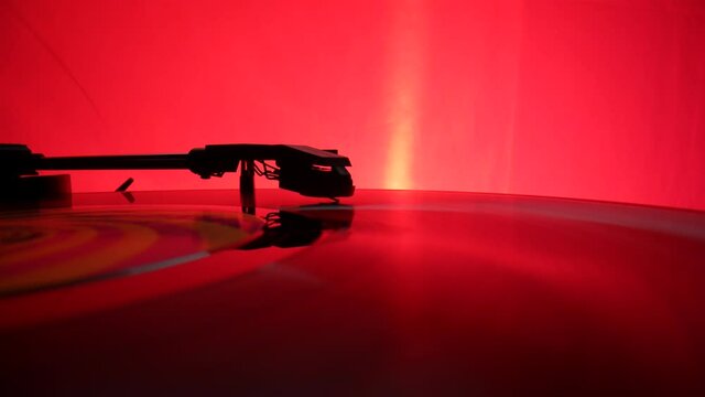 Vinyl record spins on an old turntable, illuminated by red lights. Black silhouette of turntable needle on vinyl record, reads recording from grooves on disc. Retro record vinyl player. Slow motion.
