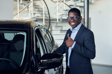 Fototapeta na wymiar Standing near car. Young african american businessman in black suit is the autosalon