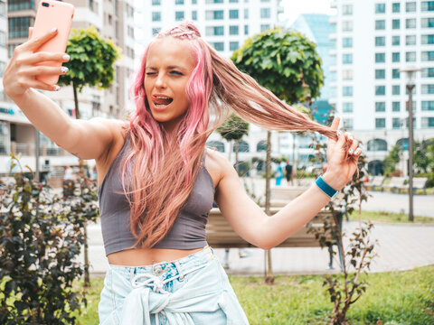 Young beautiful smiling hipster woman in trendy summer jeans shorts with pink hairstyle. Sexy carefree model posing in the street. Positive model outdoors.Taking selfie photos. Shows tongue