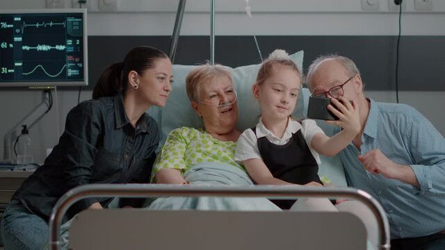 Family taking pictures with old patient in hospital ward, using mobile phone in visit. Niece and woman taking selfie on smartphone with sick pensioner and visitors. Little girl visiting grandma
