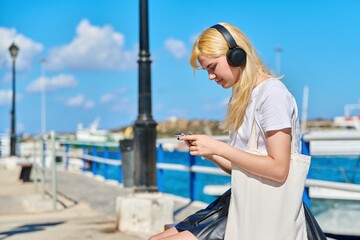 Teen female in headphones with smartphone, textile eco bag on embankment.