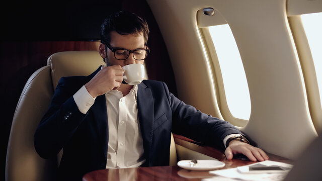 Businessman in eyeglasses drinking coffee near smartphone and papers in private plane.