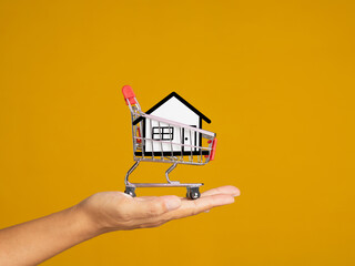 Fototapeta na wymiar Home loan and real estate investment concept. Close-up of a paper white house in the mini shopping trolley on the palm while standing on a yellow background