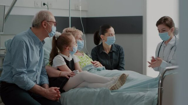Doctor explaining diagnosis to people in hospital ward during covid 19 pandemic. Physician talking about disease with patient and family in visit, wearing face mask at consultation.