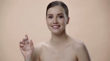 happy young woman with bare shoulders holding bottle with luxurious perfume isolated on beige.