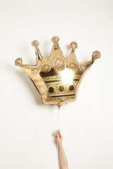 Shot of a balloon shaped as a golden crown. The golden ball is held by a hand of a person. The photo is taken on a light background.