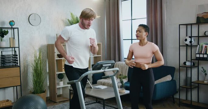 Home workout concept where attractive active sporty diverse couple training together,bearded guy running on treadmill while african american female friend supporting him