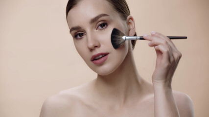 Obraz premium young woman with bare shoulders applying face powder on cheek isolated on beige.