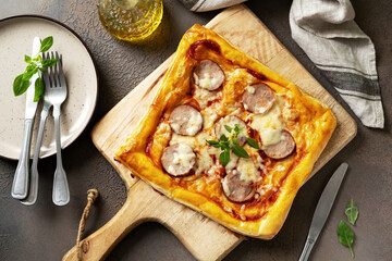 Homemade puff pastry pizza with salami, mozzarella, tomatoes and basil on the kitchen table. Delicious layered pie with sausage and cheese on a dark culinary background top view