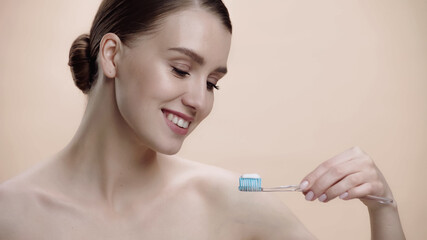 joyful woman with bare shoulders holding toothbrush with toothpaste isolated on beige.