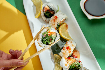 A hand holds a sushi roll with cream cheese, cucumber and salmon on bright backgrounds.