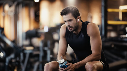 Fototapeta na wymiar tired sportsman in earphone listening music and holding sports bottle in gym.