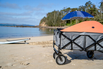 Outdoor beach cart wagon, beach umbrella an stand up paddle board on a sandy beach near the ocean. Family vacation holidays