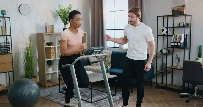 Handsome sporty muscular bearded guy supporting with applauses his active athletic fit african american girlfriend which running on treadmill at home