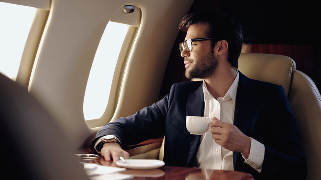 Smiling businessman holding cup and looking at window in private plane.