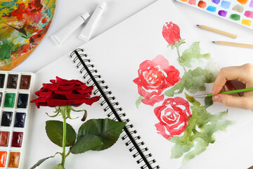 Woman painting roses in sketchbook at white table, top view