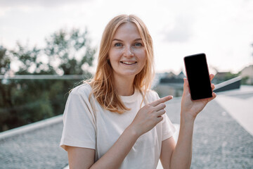 Portrait of attractive young blonde hair woman with smile, showing empty black smartphone screen, demonstrate an app or online shopping store in the park. Lifestyle concept