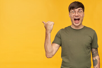 Studio shot of young tattooed brunette male, wears t-shirt and round glasses looking into camera with shocked facial expression, points with a finger at copy space. isolated over yellow background