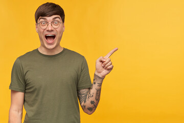 Studio shot of young tattooed brunette male, wears t-shirt and round glasses looking into camera with shocked facial expression, points with a finger at copy space. isolated over yellow background