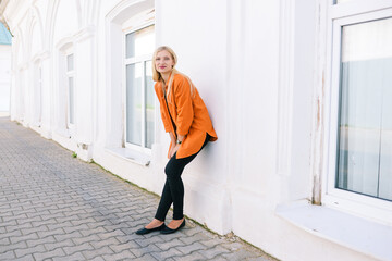 Young girl in an orange shirt and black trousers near a white wall. Happy blonde outdoors. Positive and joyful concept