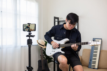 Young asian musician teaching the rock guitar class online live streaming on smartphone with student. Korean man playing electric guitar singing a song and record by cell phone shaed online social - obrazy, fototapety, plakaty