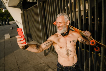 Adult shirtless man holding cellphone while standing with skateboard