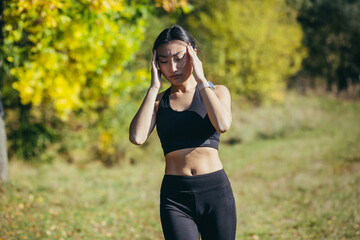 A female athlete runs in the park, has a severe headache, Asian woman holds her hands behind her head