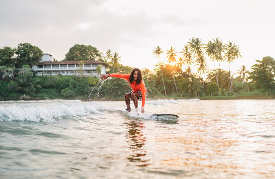 Black long-haired teen boy riding a long surfboard. He caught a  wave in an Indian ocean bay with magic sunset background. Extreme water sports and exotic countries concept.