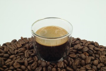 Espresso shot in a clear glass surrounded by coffee beans on a white screen.
