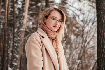 Mid age woman in finter forest looking at camera. Blonde girl in coat eyeglasses and sweater outdoors