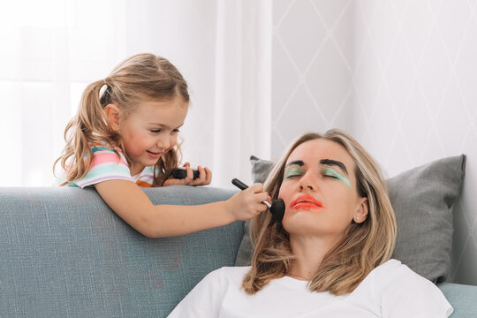 Happy loving family. Young mother sleeping on sofa at home while child girl paint face colored watercolors.Funny make up.having fun.Prank